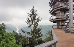Een houten terras met een metalen hek dat uitkijkt over een vallei met een rivier en verre bergen.