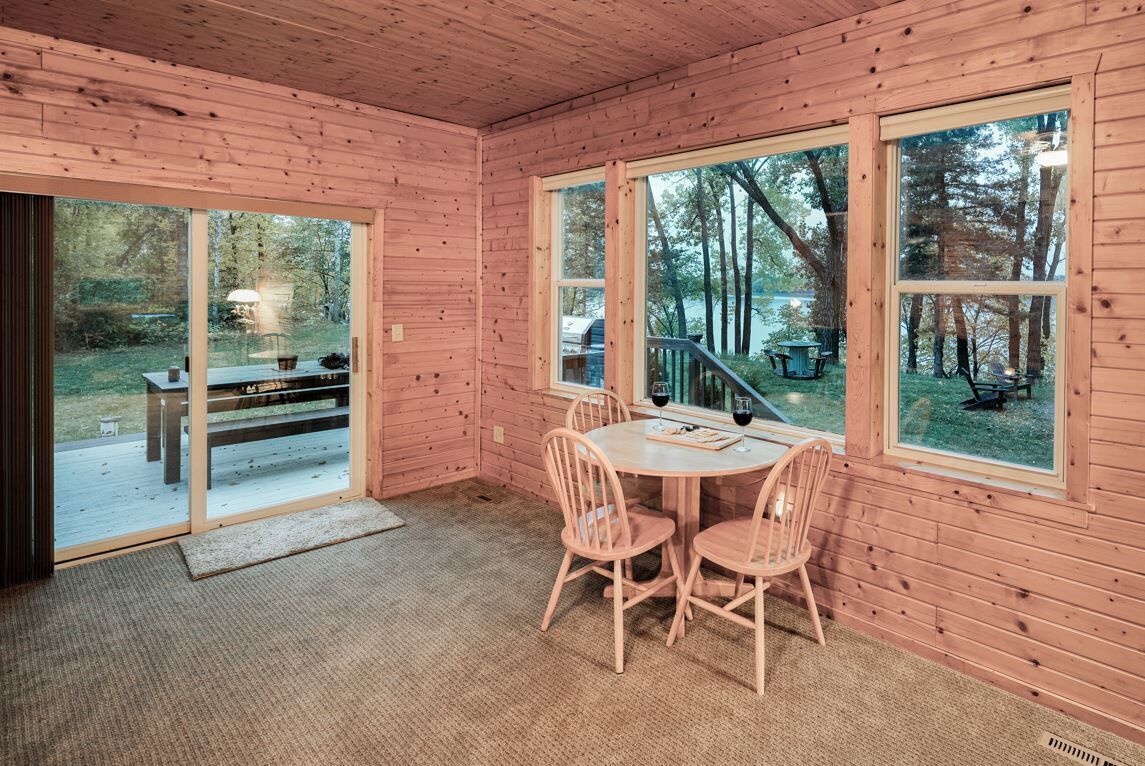 Interior de una cabaña de madera con una mesa de comedor y sillas, ventanas amplias y vistas al exterior.