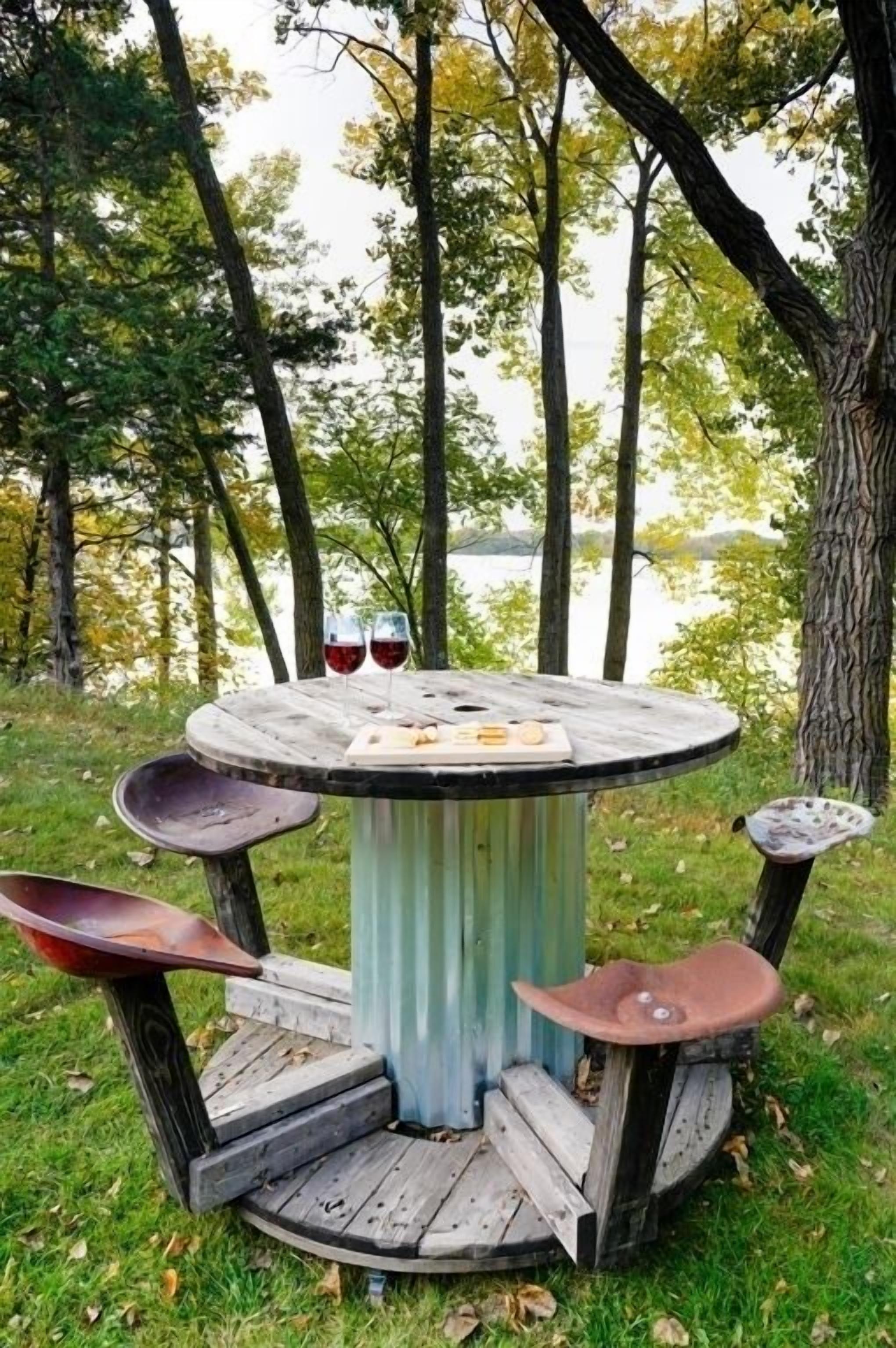 Una mesa rústica para comer al aire libre con tapa de madera y patas de metal, rodeada de un césped con árboles y una masa de agua al fondo.