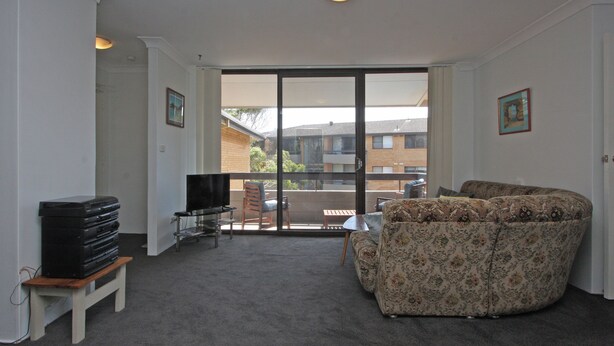 A living room with a sofa, a TV, and a small table. There is a sliding glass door leading to a balcony with a view of buildings. - Berdella 34 - Flynn's Beach (Port Macquarie)