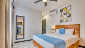 A hotel room with a wooden bed, a ceiling fan, a kitchen visible through a doorway, and a wall-mounted air conditioner.