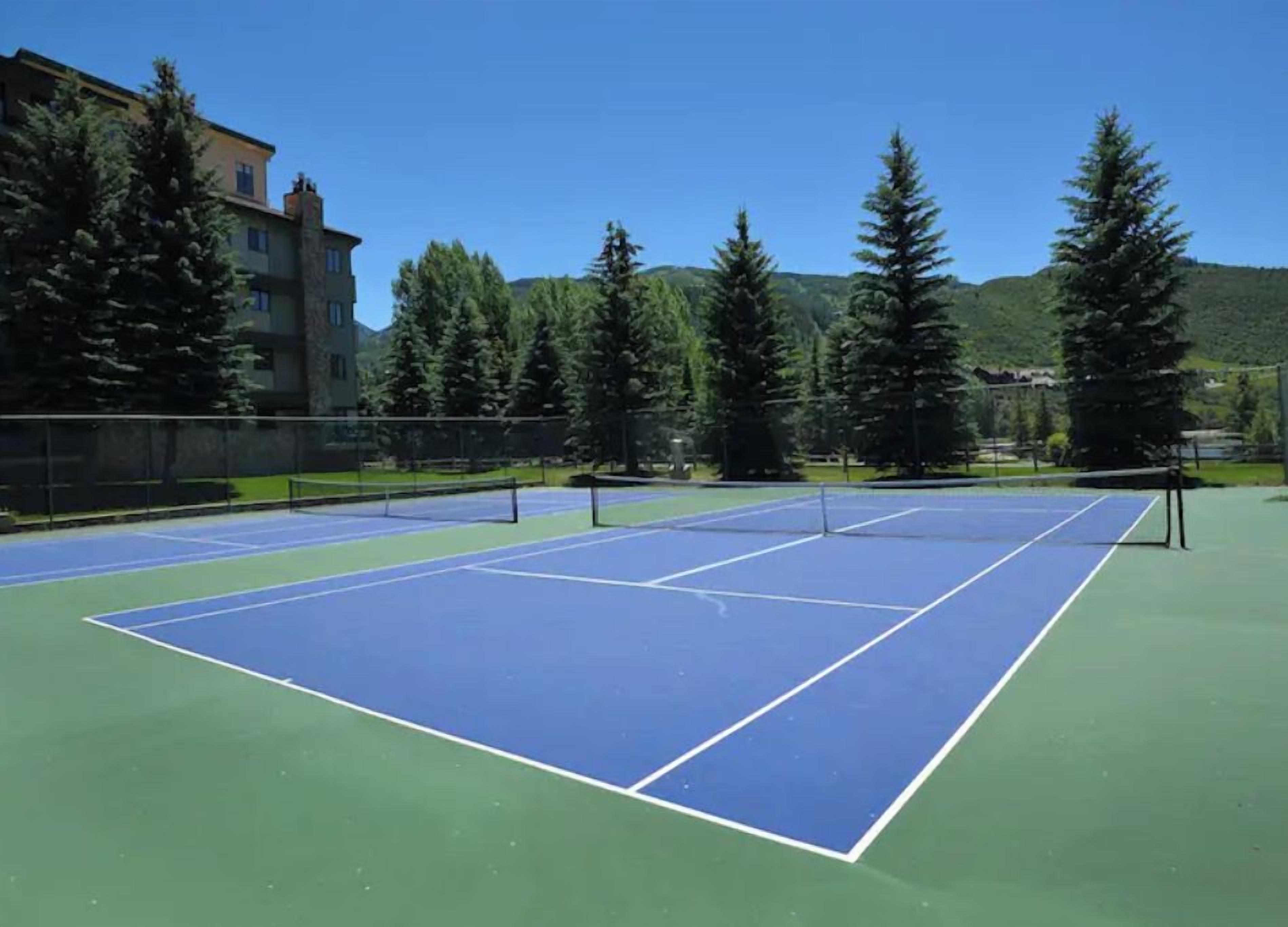 Tenniskenttä, jossa on verkko, ja jonka ympärillä on puita ja rakennuksia.