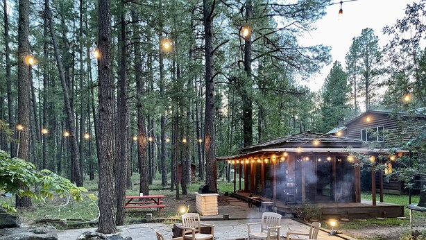 Un espace extérieur accueillant, aménagé avec un foyer, des chaises en bois et des guirlandes lumineuses, le tout dans un cadre forestier.