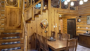 L’intérieur d’un chalet en bois comprend une table à manger, des chaises, un escalier avec des rampes en bois et un lustre.