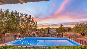 A rectangular swimming pool with a tiled deck, surrounded by a black metal fence, set against a backdrop of trees and a sunset sky. - Serene Home ~ Sleeps 16 ~ Kings & Queens~ 2 Kitchens~ Hot Tub ~ Starlink Wifi (Sedona)