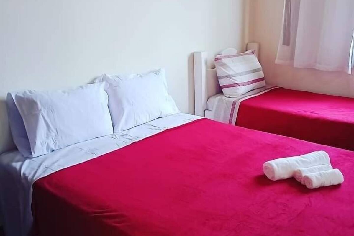 A hotel room with two beds, each with a red bedspread and white pillows. There is a window with white curtains, and a small table with a chair.