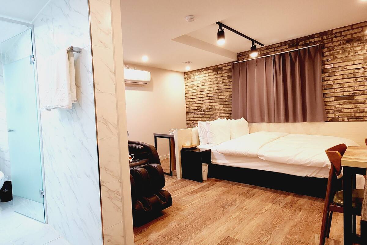 A modern hotel room with a marble bathroom, a wooden floor, a bed with white linens, a leather sofa, and a brick wall.