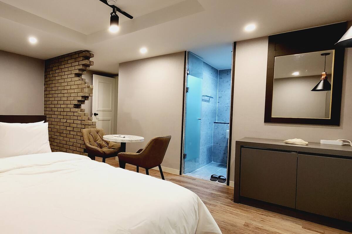 A hotel room with a large bed, a small round table, a chair, a wardrobe, and a shower area.