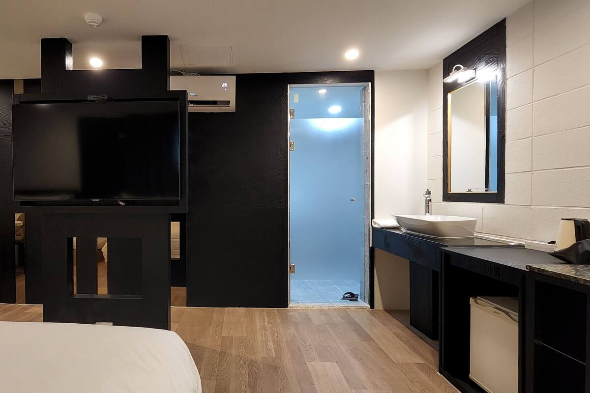 A modern hotel room with a bed, a TV, a bathroom with a sink and mirror, and a door leading to another room.