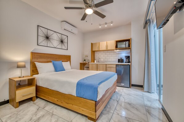 Une chambre avec un lit en bois, une table de chevet, un ventilateur de plafond et une vue sur la cuisine et le balcon.