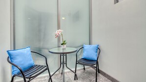 Un pequeño espacio al aire libre con dos sillas, una mesa, una planta en maceta y un panel de vidrio.