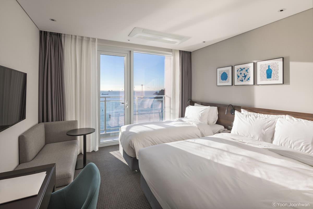 Habitación de hotel con dos camas, un escritorio, una silla y un amplio ventanal con vistas al mar.