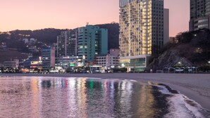 A coastal cityscape at dusk with modern high-rise buildings, a beach in the foreground, and a calm sea reflecting city lights. - Grab the Ocean SONGDO (Busan)