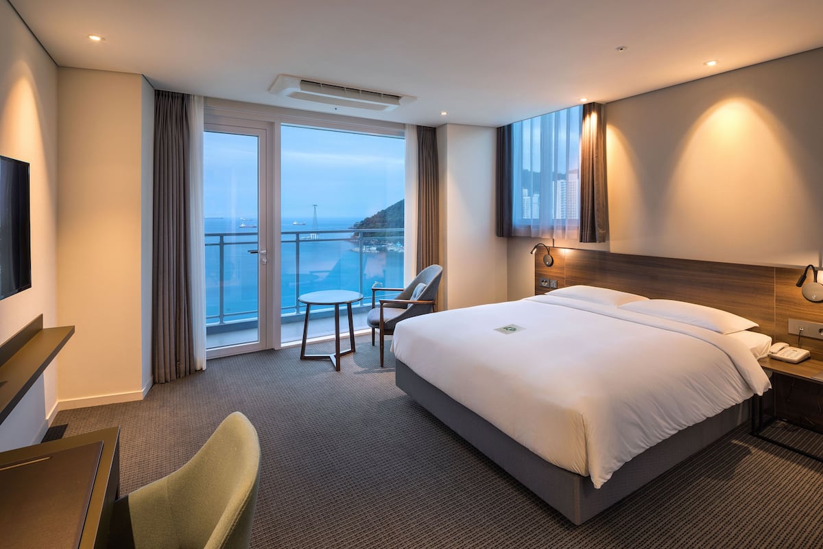 Habitación de hotel con una cama grande, un escritorio, una silla, un televisor y un balcón con vistas al mar y edificios.