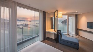 A hotel room with a large window offering a view of the sea and boats, a bed, a seating area with a table, and a TV mounted on the wall. - Grab the Ocean SONGDO (Busan)