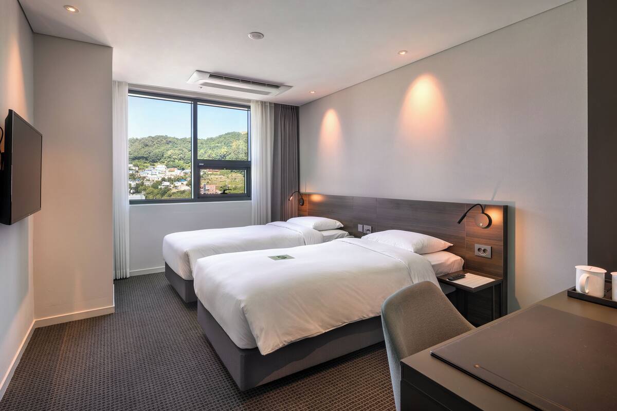 Habitación de hotel con dos camas, un escritorio, una silla, un televisor y una ventana con vistas a una ciudad y colinas.