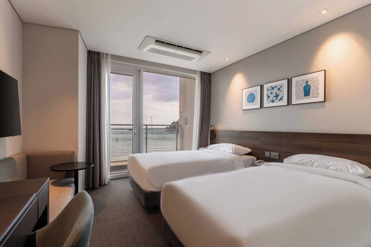 Habitación de hotel con dos camas, un escritorio, una silla, una mesita pequeña y un ventanal con vistas al mar.