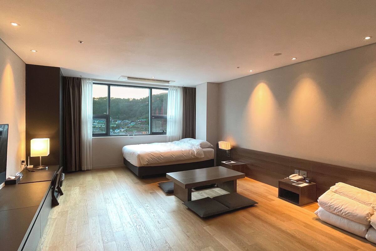 Una habitación de hotel moderna con una cama grande, un escritorio, una silla, una mesita de noche y vistas al exterior.