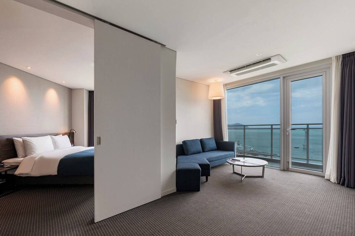 Una habitación de hotel moderna con cama, sofá, mesa de centro y balcón con vista al mar.
