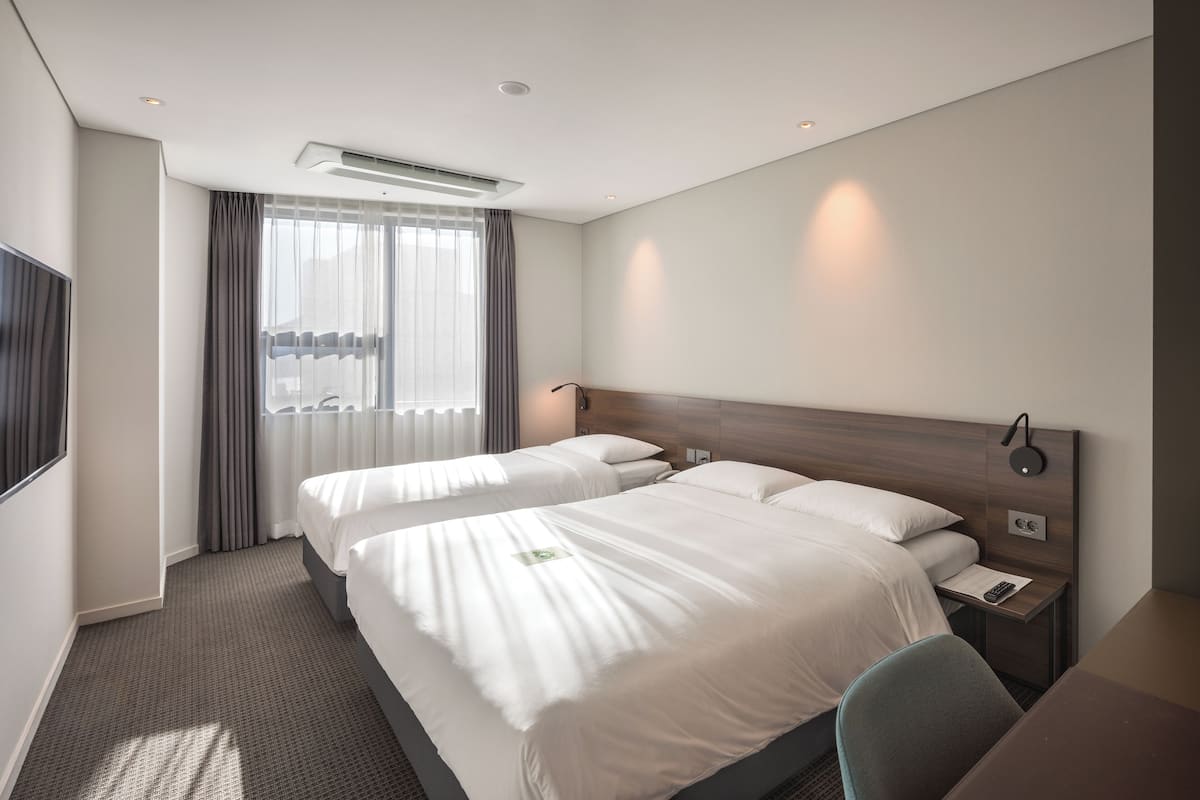 Habitación de hotel con dos camas, un escritorio, una silla, una mesita de noche, televisión y ventana con cortinas.