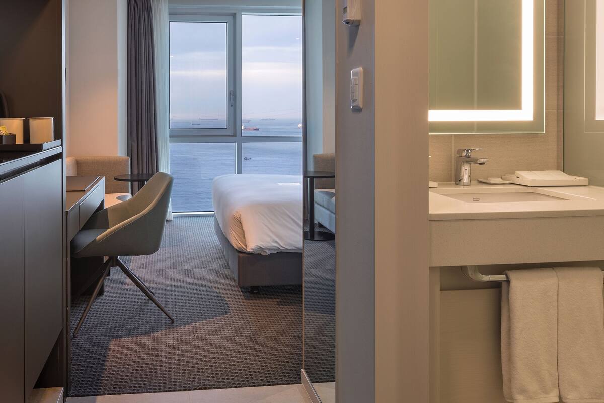 Una habitación de hotel moderna con una cama, un escritorio, una silla, un baño con espejo y vistas al mar a través de amplios ventanales.