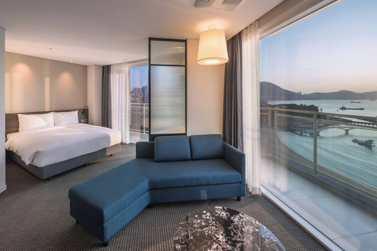 Habitación de hotel moderna con una cama grande, un sofá, vista a un cuerpo de agua y un paisaje urbano al fondo.