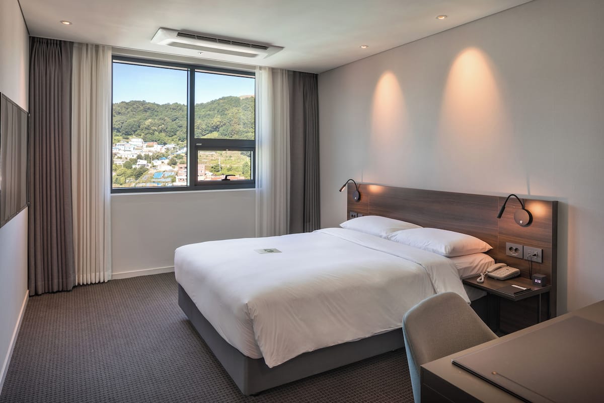 Habitación de hotel con una cama grande, un escritorio, una silla, una mesita de noche con un teléfono y vistas a un paisaje urbano a través de una ventana.