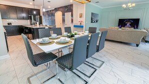 A modern kitchen with dark cabinets, a dining area with a white table set for six, and a living room with a sofa and TV. -  6 Br/5 Ba Pool/Game /Themed Handpainting Villa (Kissimmee)