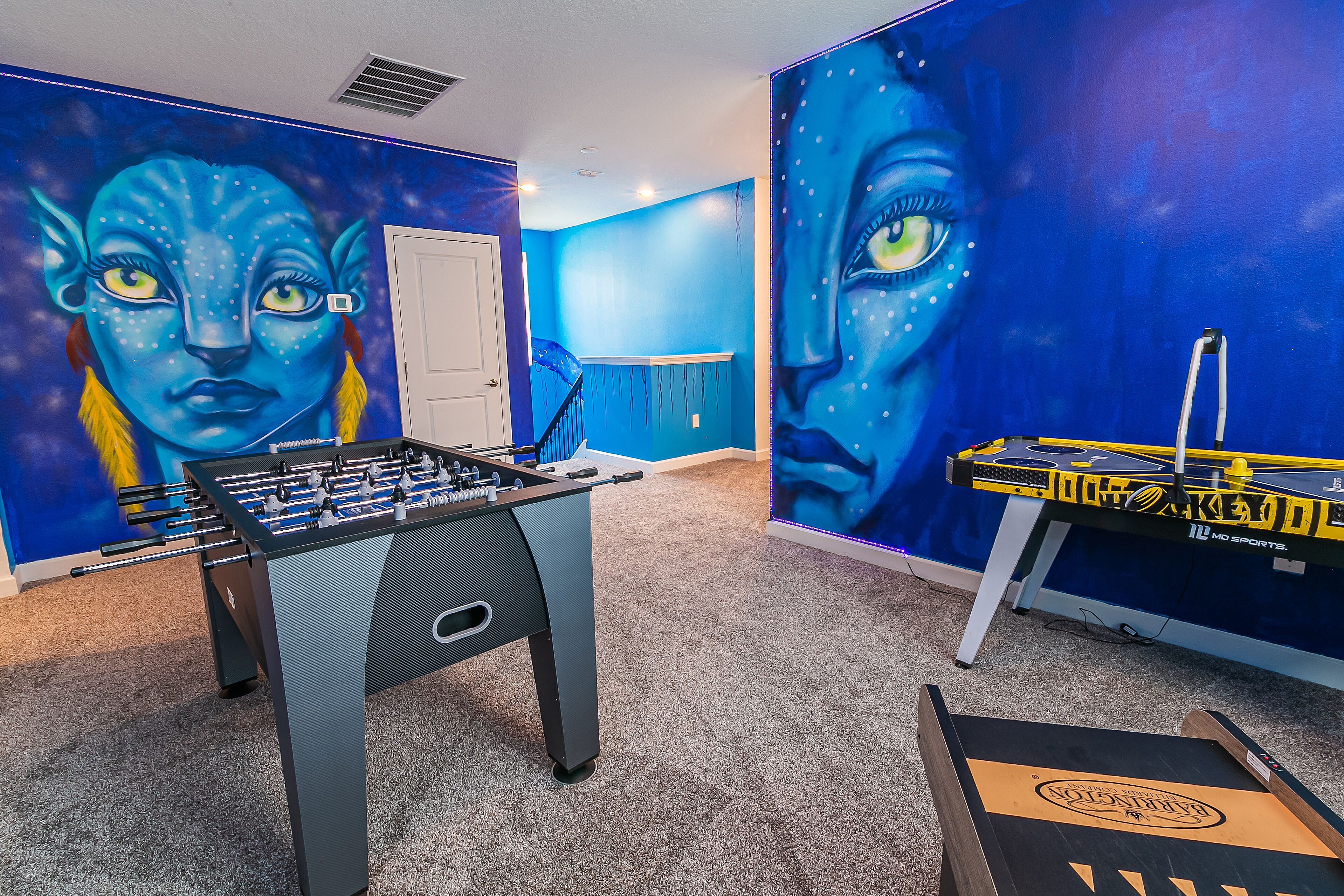 Een kamer met twee tafelvoetbaltafels, waarvan er een blauw en zwart is en de andere geel en zwart. De muren zijn blauw met een groot muurschildering van een gezicht.