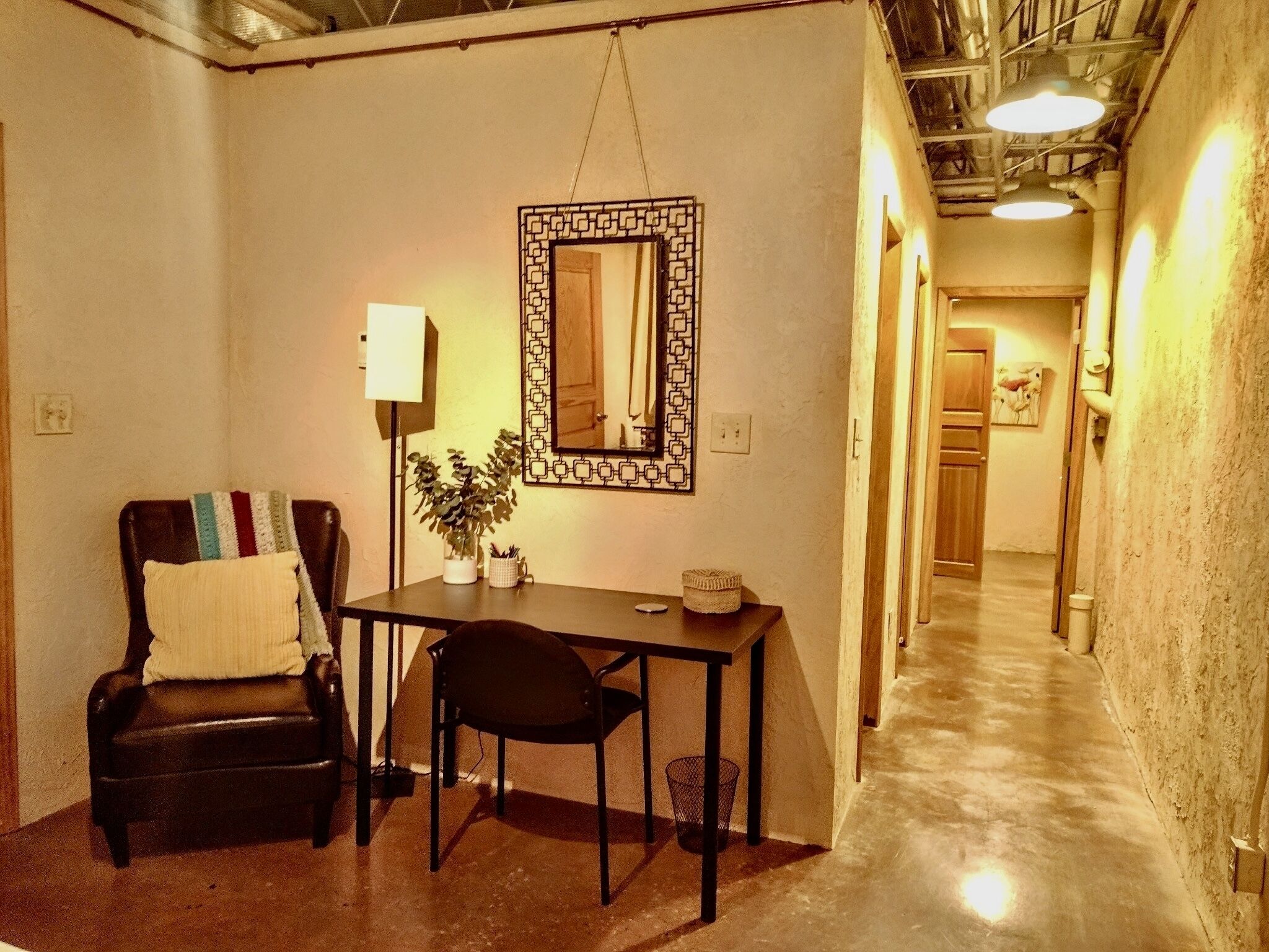 Un couloir étroit avec un plancher en bois, une petite table avec une chaise, une plante en pot et un miroir fixé au mur.