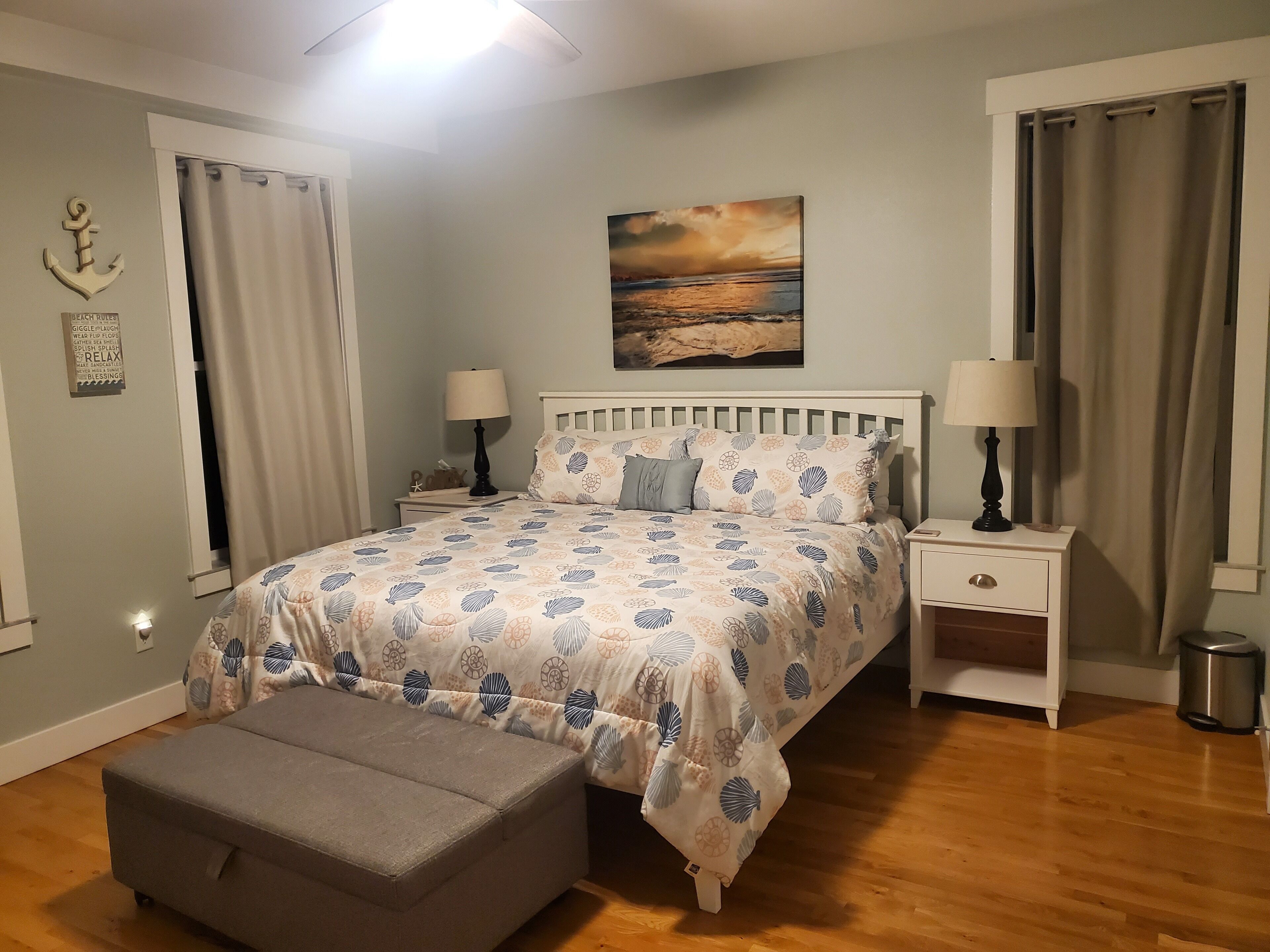 Un dormitorio con cama, dos mesitas de noche, una pequeña consola, un banco, un cuadro y cortinas.