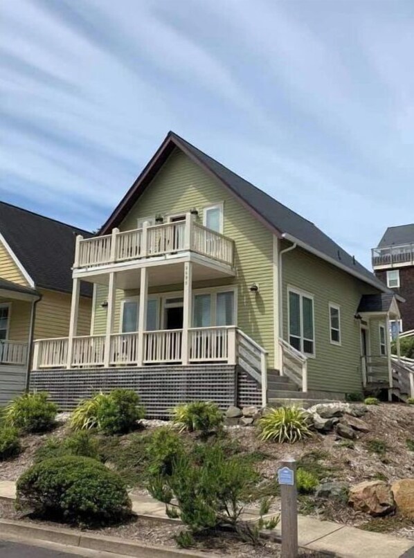 A two-story house with a balcony on the second floor, a front porch, and a small garden with shrubs and rocks. - Olivia Beach Parkside house with hot tub and short walk to beach (Lincoln City)
