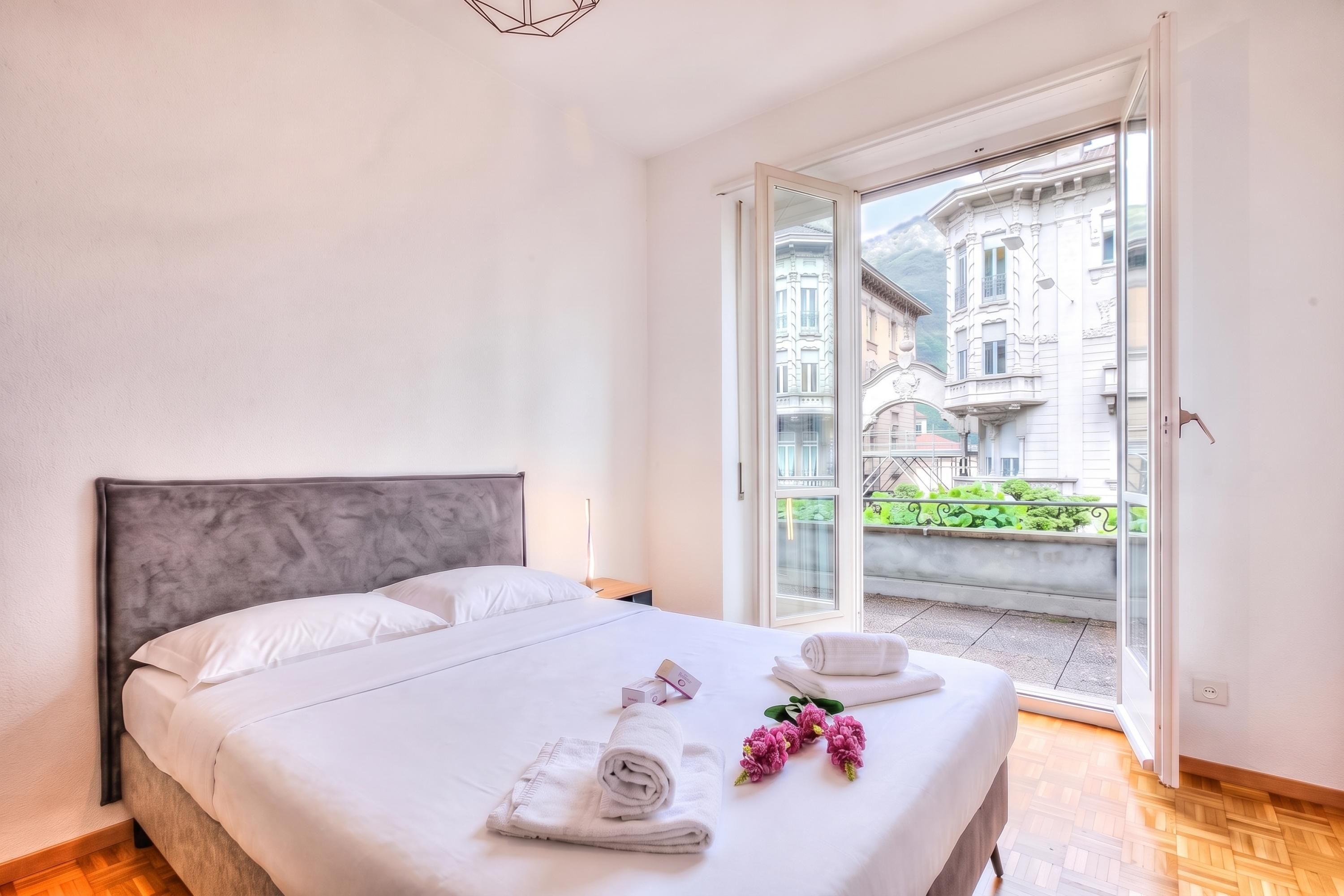 A hotel room with a bed, a nightstand, a window with a view of buildings, and a door leading to an outdoor area.