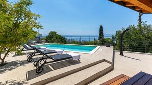 Un espace piscine aménagé avec des chaises longues et un banc en bois, offrant une vue sur une étendue d’eau et des bâtiments situés au loin.