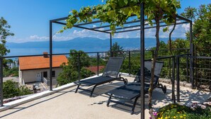 Un balcon avec deux chaises longues, une pergola couverte de plantes grimpantes et une vue sur la ville et les montagnes.