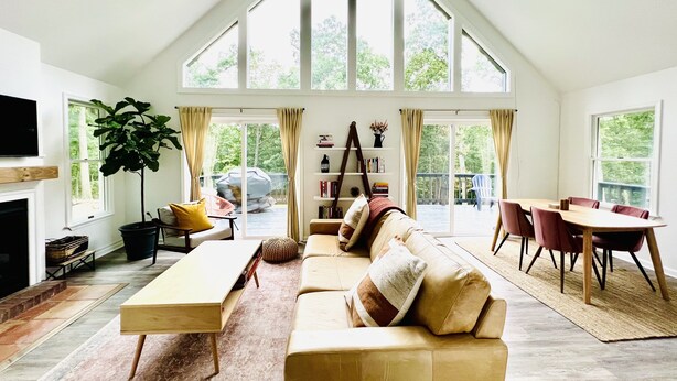 A spacious living room with a high ceiling, large windows, a wooden sofa, a dining table with chairs, and a fireplace.