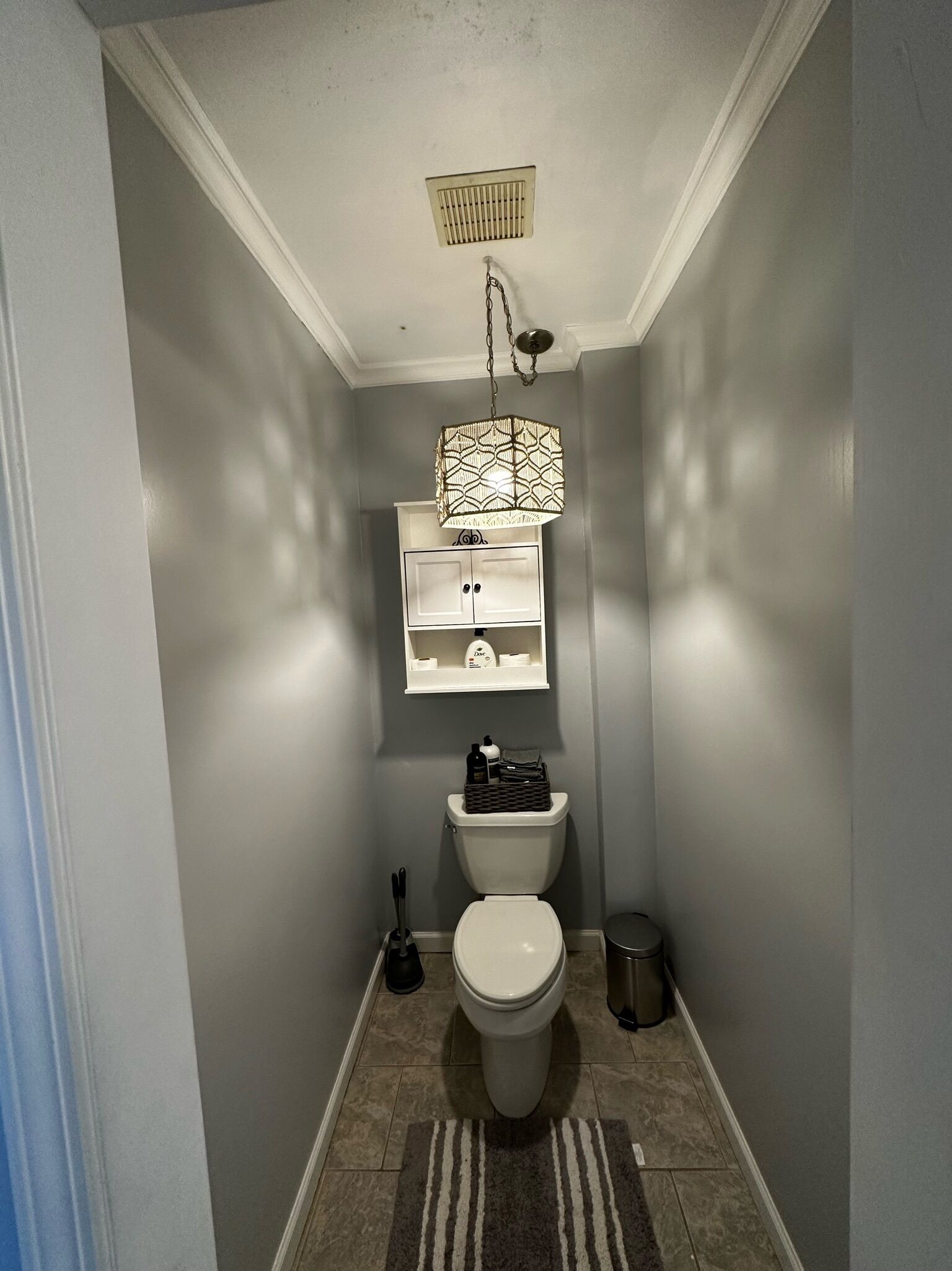 Et lille badeværelse med toilet, en hylde over toilettet, en hængende lampe og en skraldespand.