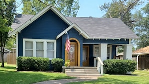 A single-story house with a dark blue exterior, a front porch, a small front yard, and an American flag.