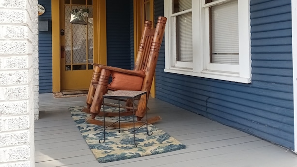 A porch with a wooden chair and table, a welcome sign, and a window with blinds.