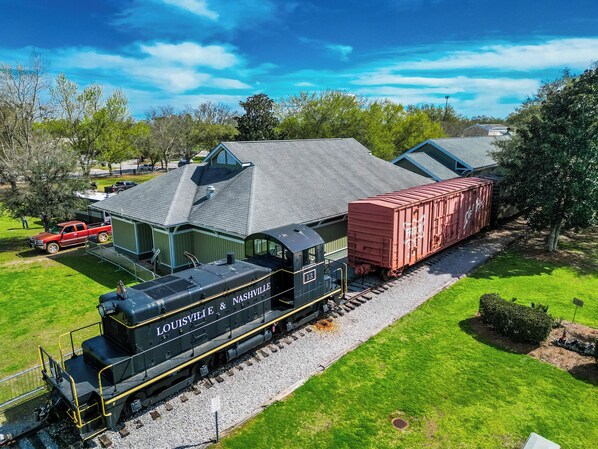 A train with "Louisville & Nashville" on the front, passing by a building with a green roof and a red truck parked nearby.