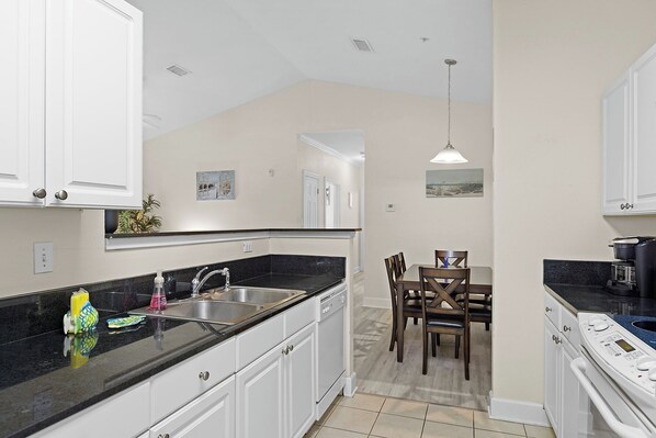 A kitchen with white cabinets, a black countertop, and a dining area with wooden chairs. - Near Tropic Falls Water Park – Fun & Relaxation Await! (Foley)