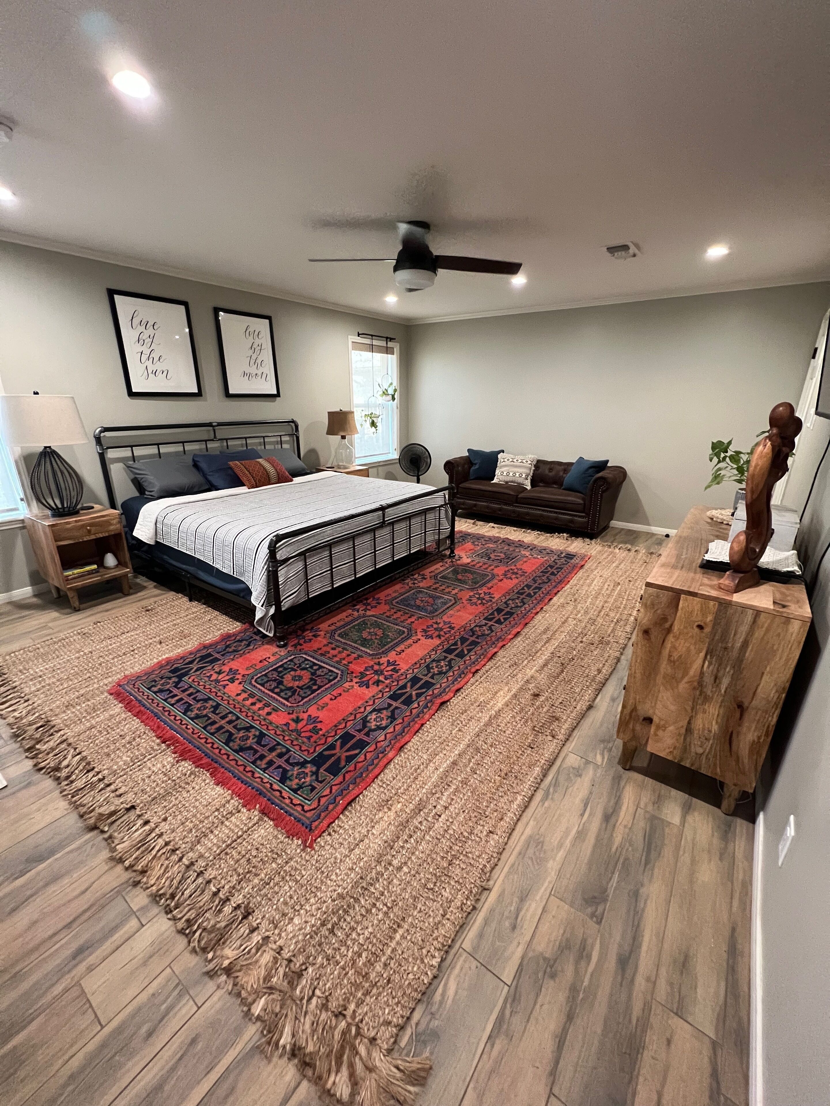A bedroom with a bed, a wooden side table, a wooden bench, a red and black rug, a ceiling fan, and two wall-mounted lights.