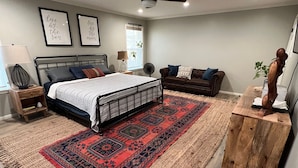 Un dormitorio con cama, una mesita de noche de madera, un banco de madera, un tapete rojo y negro, un ventilador de techo y dos luces fijadas en la pared.