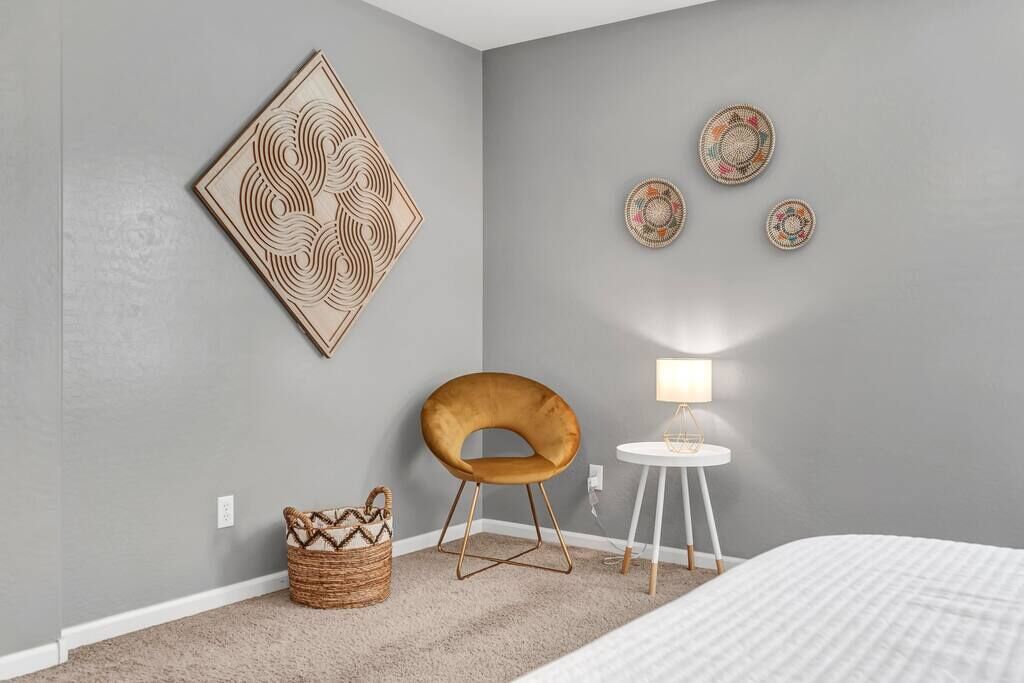 A bedroom with a grey wall, a large geometric artwork, a small round table with a lamp, a brown chair, and a woven basket.