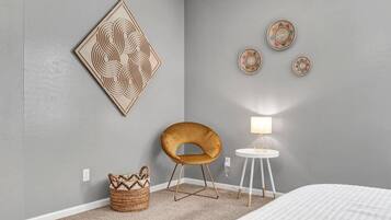 A bedroom with a grey wall, a large geometric artwork, a small round table with a lamp, a brown chair, and a woven basket.