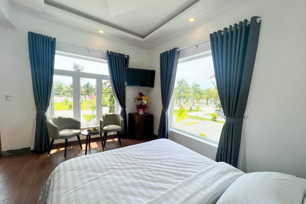 A hotel room with a bed, two chairs, a small table, a TV, and large windows with curtains.