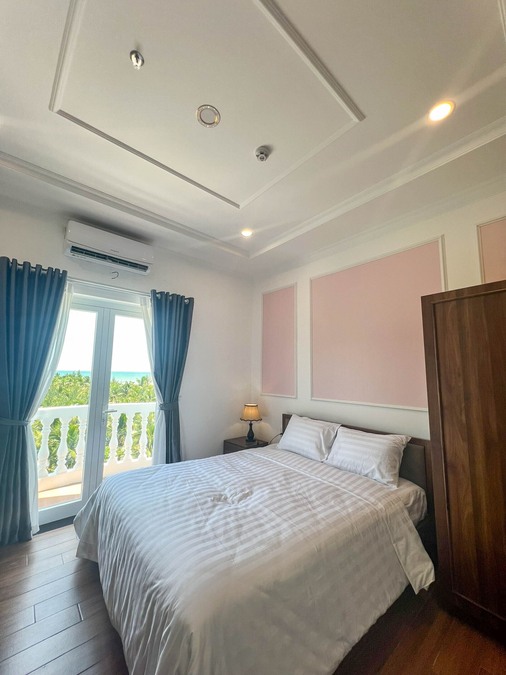 Un dormitorio con cama, mesita de noche, lámpara, ventana con cortinas y vista al exterior.