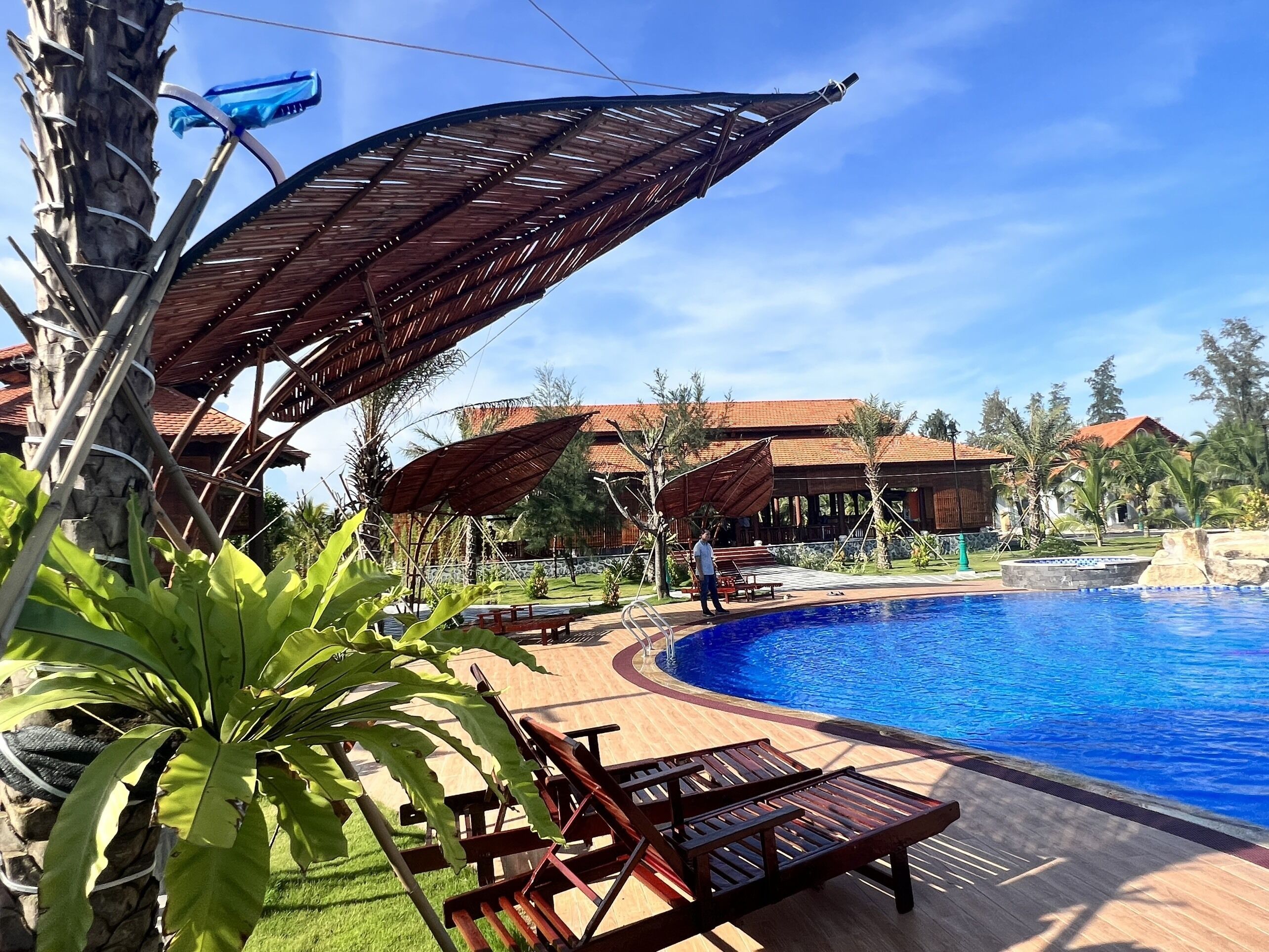 A pool area with lounge chairs and umbrellas, surrounded by greenery and a building with a tiled roof.