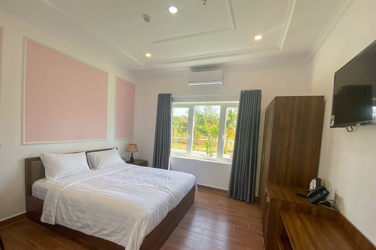A hotel room with a large bed, a wooden desk, a flat-screen TV, and a window with curtains.