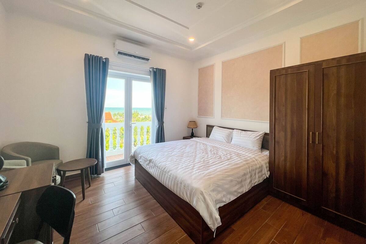 A hotel room with a large bed, a wardrobe, a desk with a chair, a small table, and a view of the sea through the balcony.